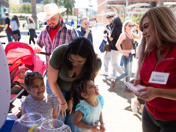 woman with a name badge talks to two young girls laughing with their mother, father in a cowboy hat and baby in a stroller
