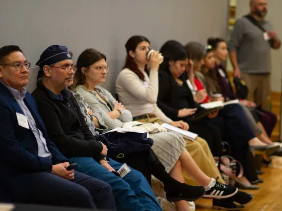 a group of people sit in chairs against a grey wall and listen to speakers