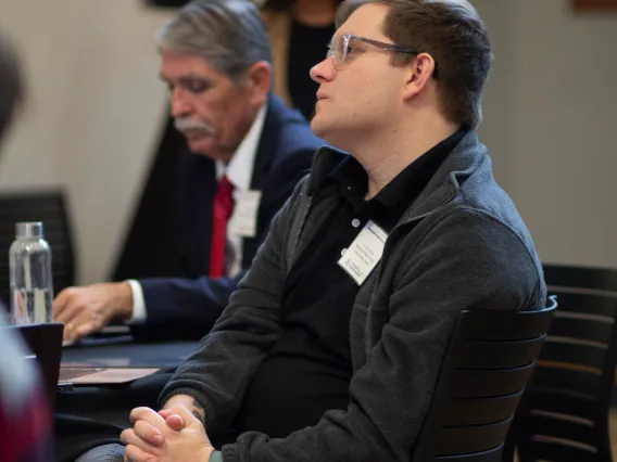 two men sit at a table listening to a speaker