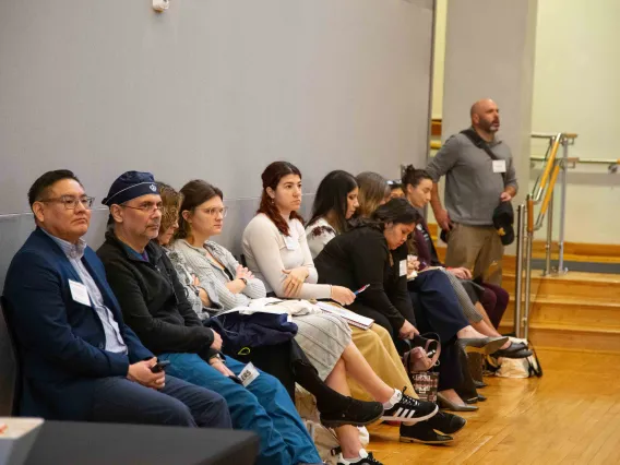 a group of people sit in a line of chairs against a grey wall listening to a speaker