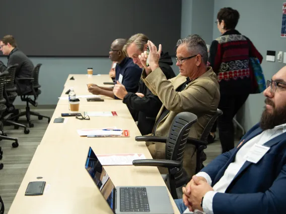 a man sitting at a long table with others holds up his smartphone to take a picture of a slide presentation