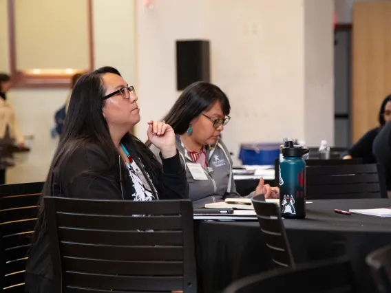 two women sit at a round table listening to a speaker