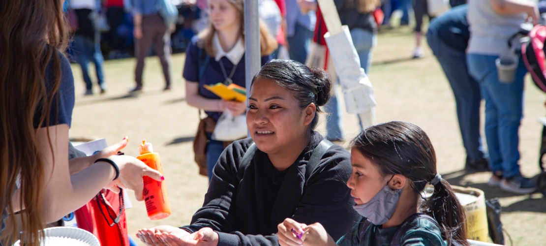 Mother and daughter sit at an activity table with hands held out blurred crowd in the background