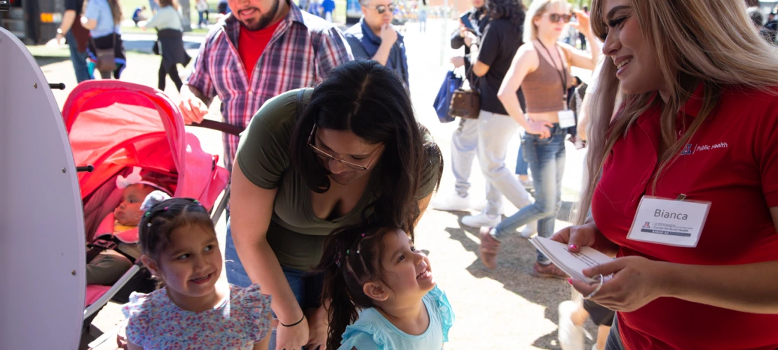 woman with a name badge talks to two young girls laughing with their mother, father in a cowboy hat and baby in a stroller