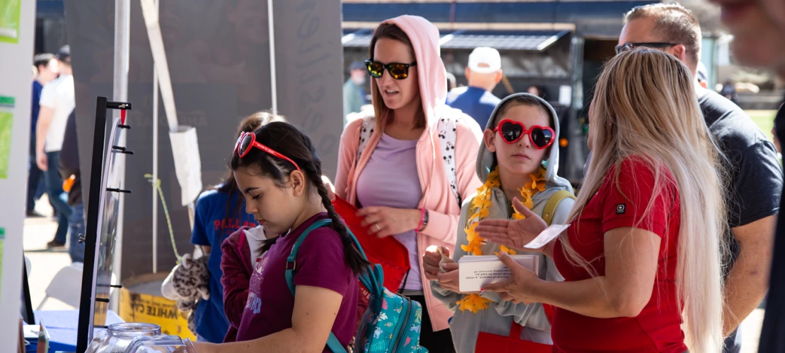 woman with long hair and a red shirt talks to a group of girls wearing heart shaped sunglasses while a girl picks out stickers from a table
