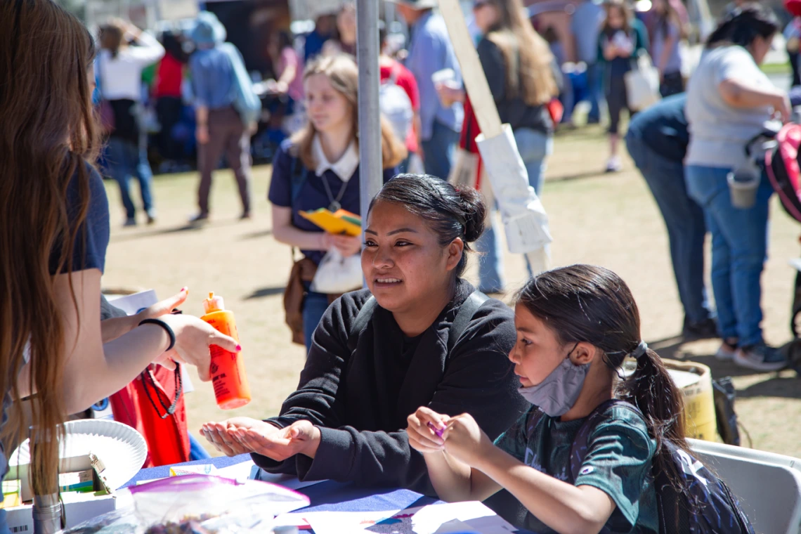 Mother and daughter sit at an activity table with hands held out blurred crowd in the background