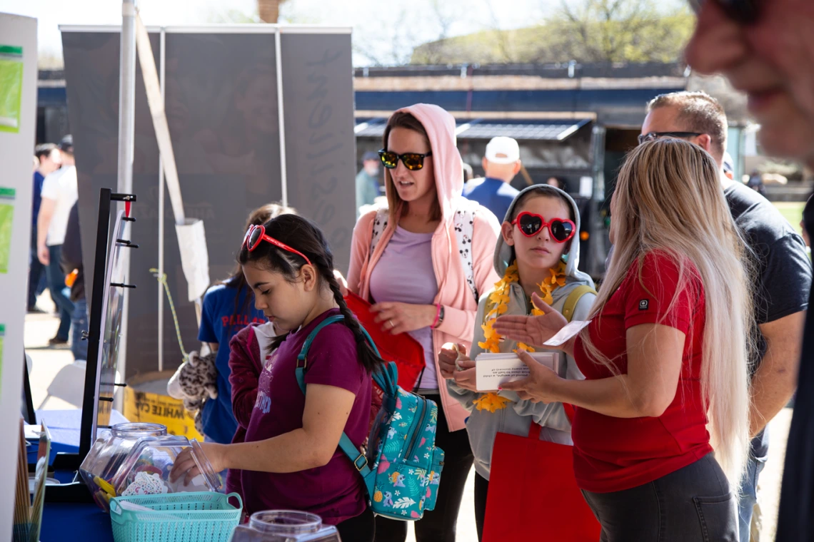 woman with long hair and a red shirt talks to a group of girls wearing heart shaped sunglasses while a girl picks out stickers from a table