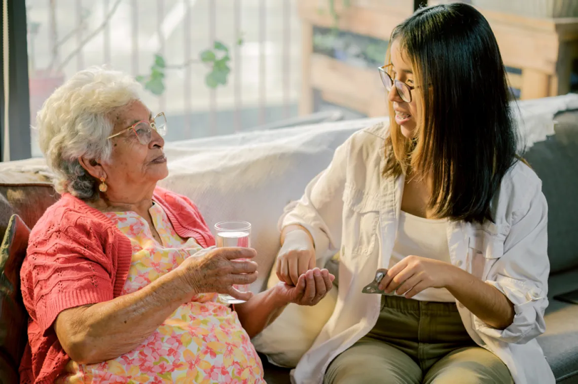 a young woman with brown hair and glasses sits with an older woman holding a glass of water