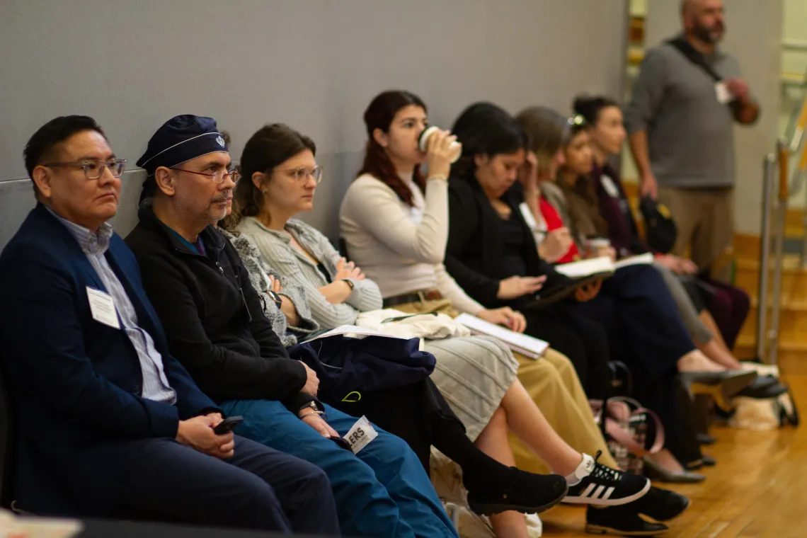 a group of people sit in chairs against a grey wall and listen to speakers