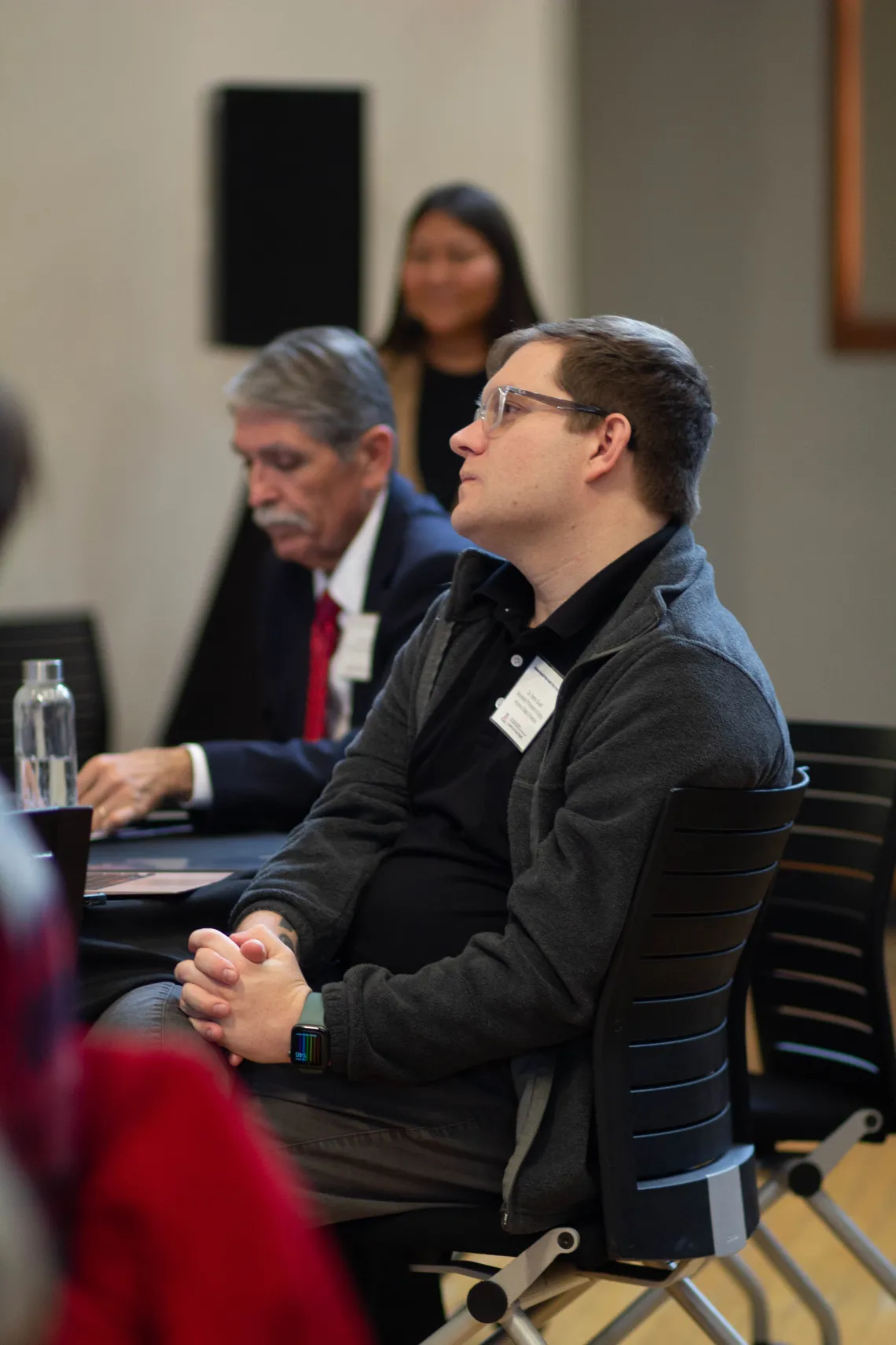 two men sit at a table listening to a speaker