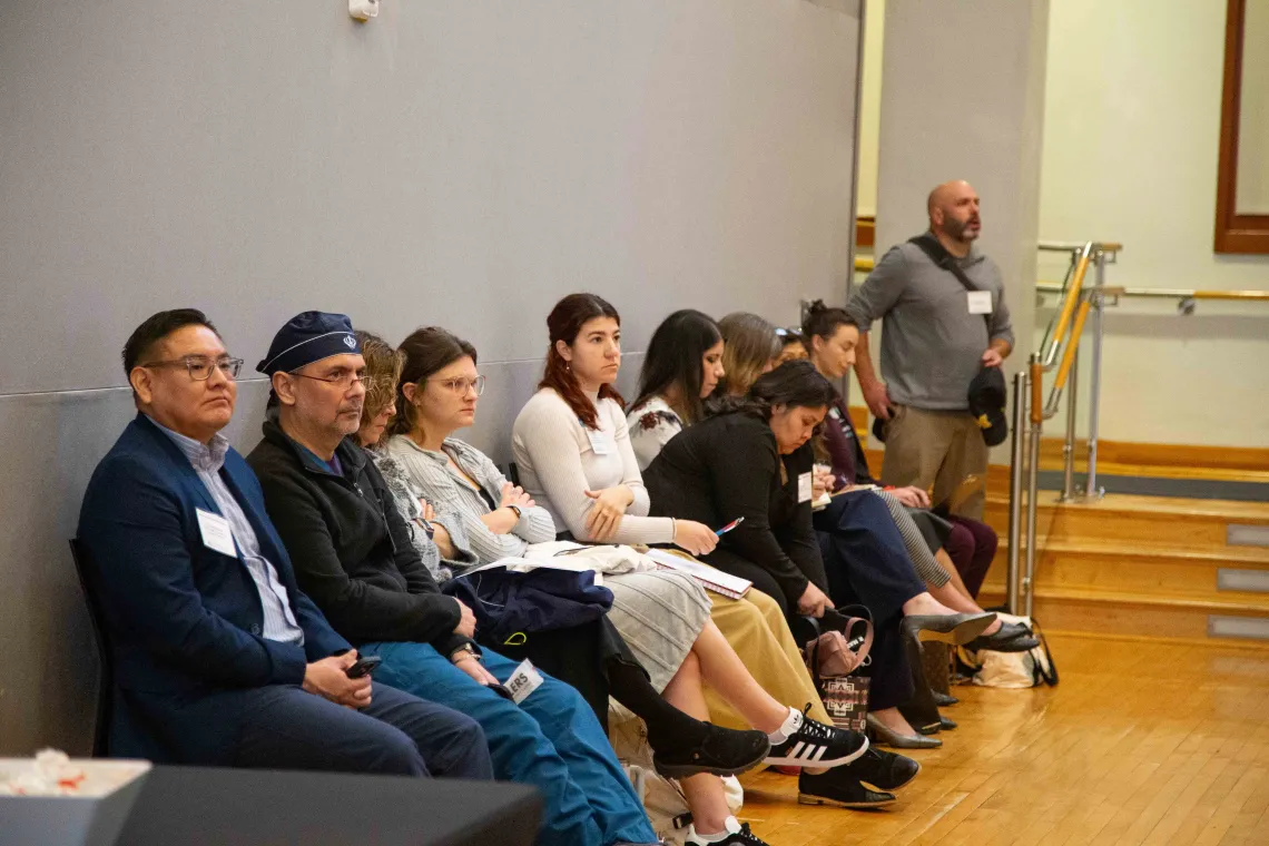 a group of people sit in a line of chairs against a grey wall listening to a speaker