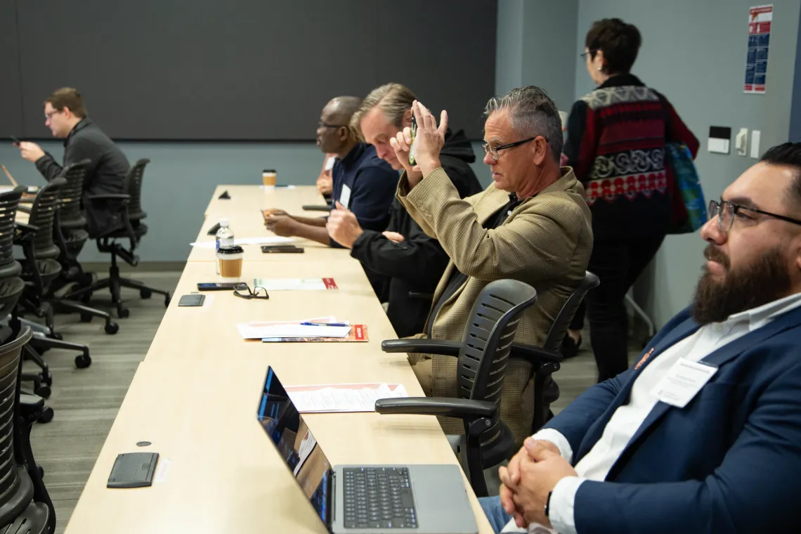 a man sitting at a long table with others holds up his smartphone to take a picture of a slide presentation