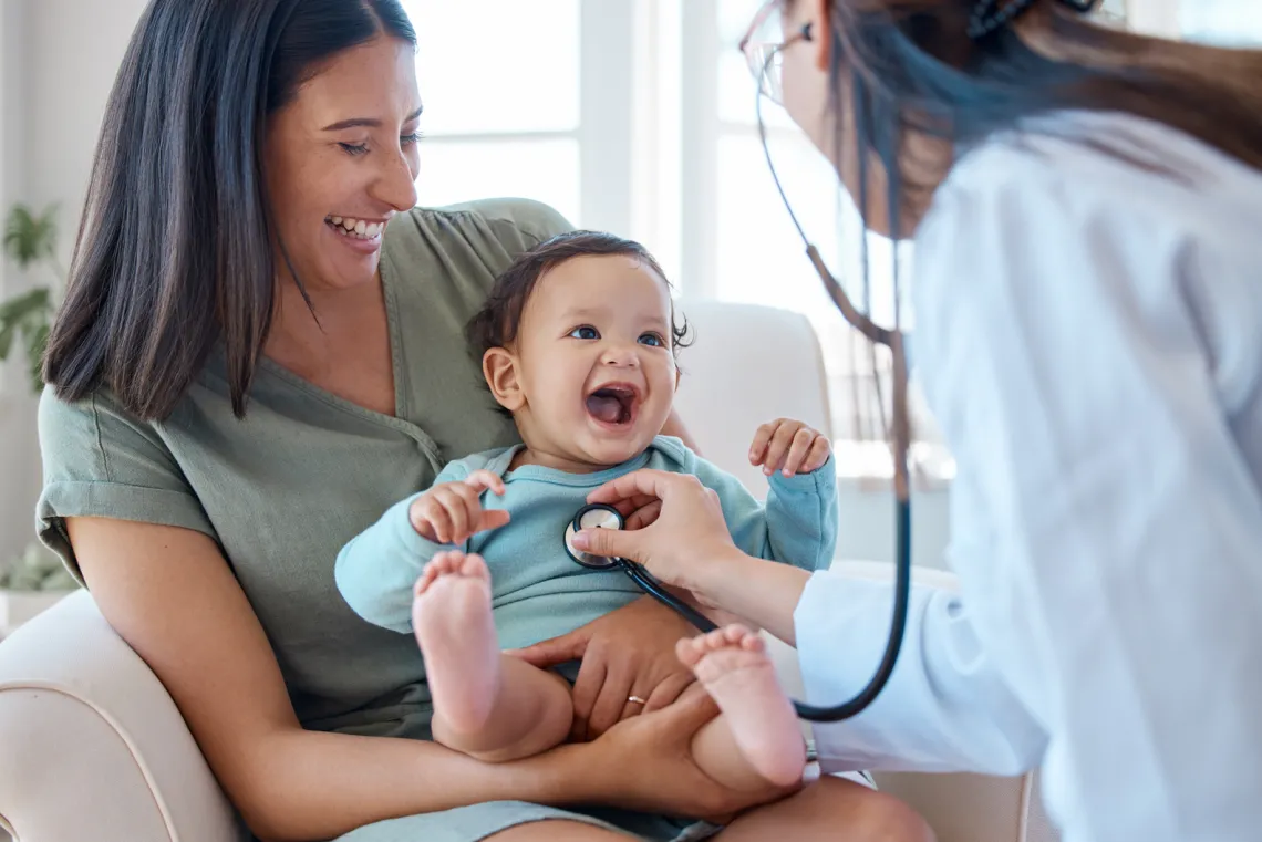 baby laughs on their mother's lap while doctor listens with stethoscope