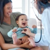 Baby laughing in mothers lap while doctor listens with stethoscope